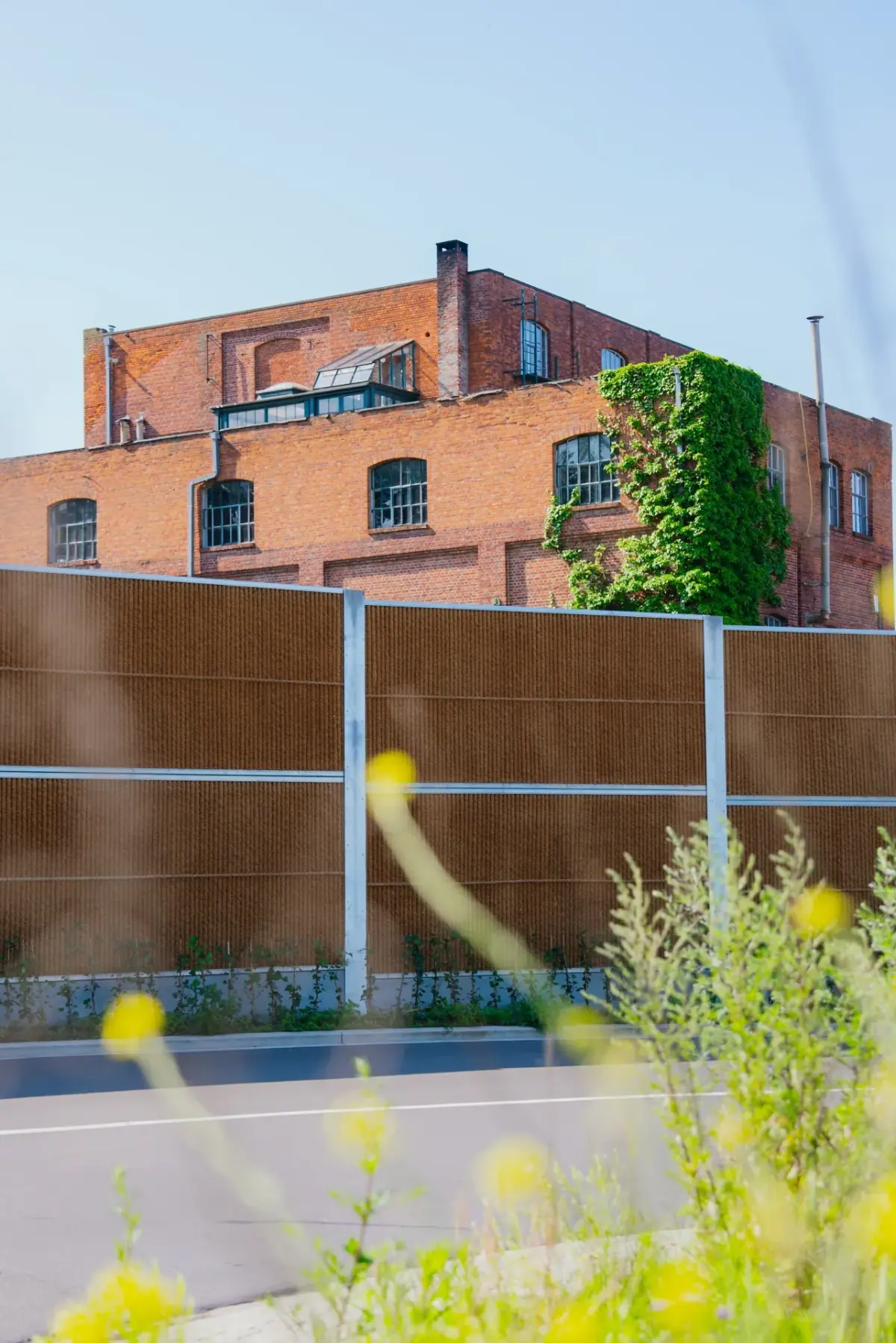 Kokowall geluidsscherm langs een weg met natuurlijke kokosvezel afwerking en bakstenen industriegebouw op de achtergrond